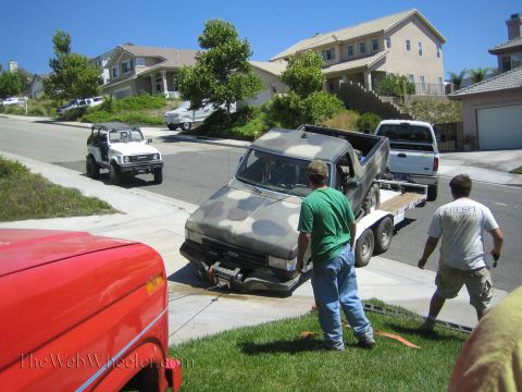 1990 Ford Bronco - Two men maneuver a camouflage-painted Ford vehicle body off a flatbed trailer in a suburban driveway, with a white Suzuki 4x4 parked at the…