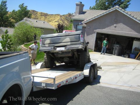 1990 Ford Bronco - lifted camo-painted truck rests on a tandem-axle trailer in a driveway as two men work nearby outside an open garage.
