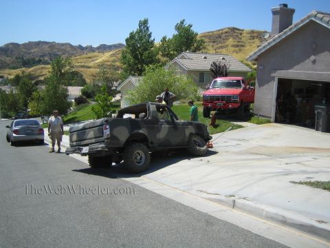 1990 Ford Bronco - lifted camouflage-painted pickup with its cab opened up sits in a driveway while men work, with a red lifted Ford truck behind it.