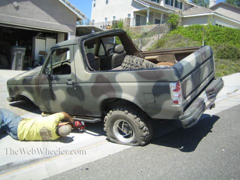 1990 Ford Bronco - man lies beneath a camouflage-painted Ford SUV with its top removed, parked along a curb in front of suburban homes.