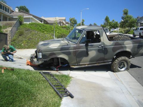 1990 Ford Bronco - man kneels at the front of a camouflage-painted Ford SUV that has its front wheels removed, parked on a driveway with metal ramps laid out.
