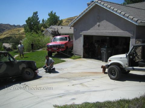 1990 Ford Bronco - camo-painted truck, a lifted red Ford pickup, and a white Suzuki 4x4 are arranged around a driveway as a man crouches and works.