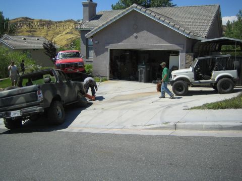 1990 Ford Bronco - Men work on a camouflage-painted truck in a suburban driveway alongside a red Ford pickup and a white Suzuki 4x4 near an open garage.