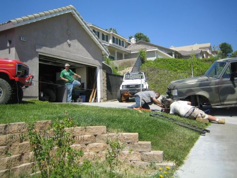 1990 Ford Bronco - Several men work on a camouflage-painted SUV in a driveway with ramps laid out, a white Suzuki and a red truck nearby.