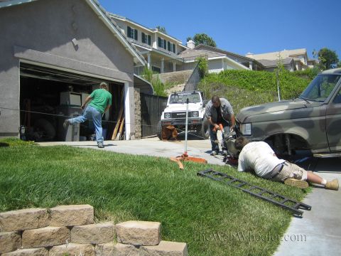 1990 Ford Bronco - Men crouch over tools and metal ramps while servicing a camouflage-painted SUV in a driveway, with a white Suzuki parked behind.