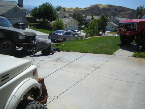 1990 Ford Bronco - person kneels at the front of a camo-painted truck holding a winch line stretched across a driveway toward a red lifted Ford truck.