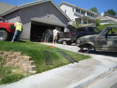 1990 Ford Bronco - camouflage-painted truck is raised on ramps and a jack in a driveway as men work near the garage, with red and dark trucks nearby.