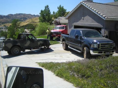 1990 Ford Bronco - blue Ford crew-cab pickup is parked in a driveway alongside a camouflage-painted truck and a red Ford pickup in front of a house.