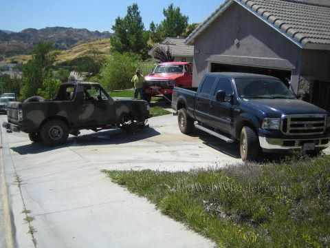 1990 Ford Bronco - blue Ford pickup and a camouflage-painted truck sit linked by a line in a driveway, with a red Ford and a man in the background.