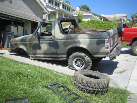 1990 Ford Bronco - Side view of a camouflage-painted Ford SUV with its top removed in a driveway, with large off-road tires and ramps lying on the grass.