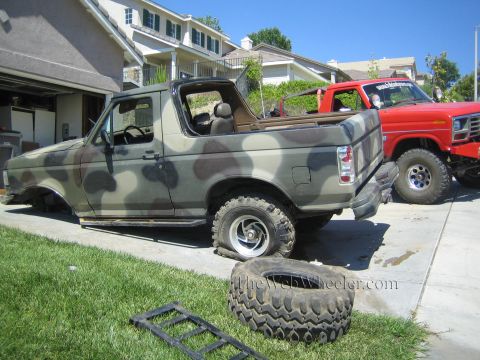 1990 Ford Bronco - camouflage-painted Ford SUV with its top removed sits in a driveway next to a red Ford truck, with spare tires and ramps on the grass.