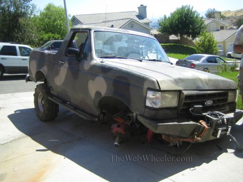 1990 Ford Bronco - camouflage-painted Ford pickup sits in a driveway with its front wheels removed and a winch and jack visible at the exposed front axle.