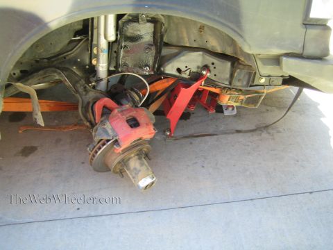 1990 Ford Bronco - Close-up under a fender showing a vehicle's front suspension, brake rotor and caliper, and a red floor jack supporting the assembly.