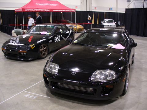 1995 Ford Taurus - Two sports cars are displayed indoors under a red tent with event branding.