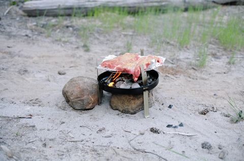 1979 Ford Bronco - Meat is grilled over coals on a portable grill outdoors, with rocks providing support on sandy terrain.