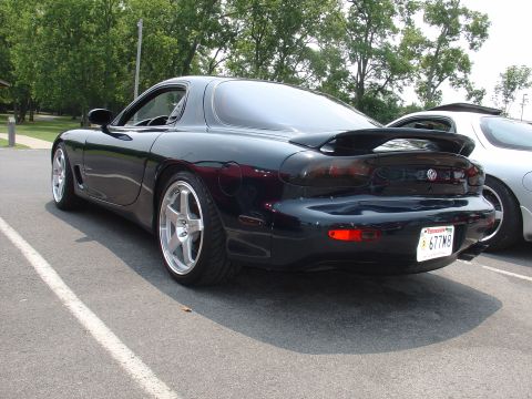 1986 Mazda RX-7 - A dark blue Mazda RX-7 is seen from the rear, parked next to another vehicle. The car features aftermarket wheels and a prominent rear spoi…