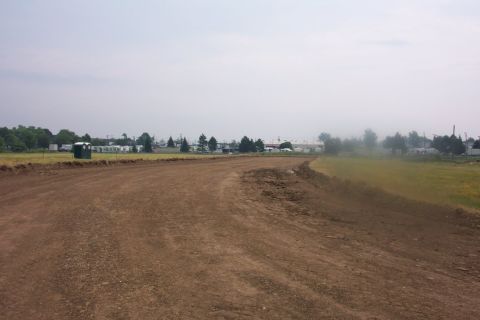 1978 Ford Bronco - A dirt track curves through a field, with a portable toilet and distant buildings visible under an overcast sky.