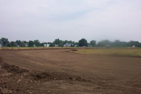 1978 Ford Bronco - A wide shot of a dirt field with houses and trees visible in the distance under a cloudy sky.