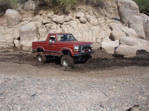 1984 Ford Bronco - This red SUV is navigating a muddy, rocky terrain with its tires deep in the muck.