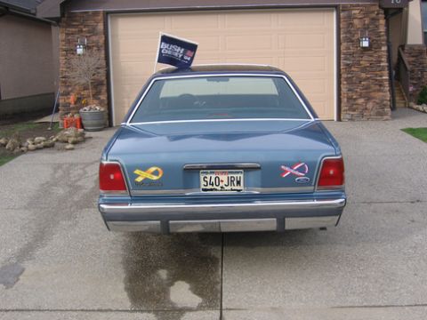 1988 Ford Crown Victoria LTD - Rear view of a blue sedan with a political flag and various stickers.