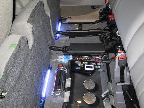 Under-seat custom car audio setup with clear capacitor, blue neon lights, and wiring in an SUV interior.