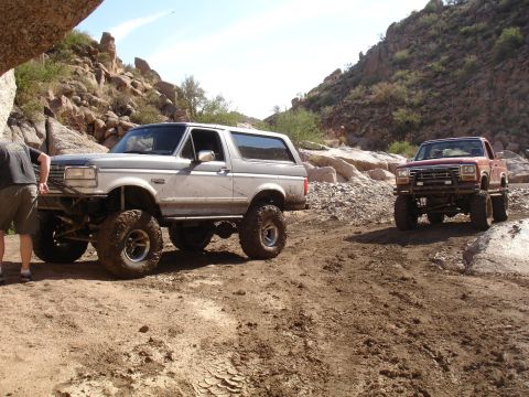 1984 Ford Bronco - Two off-road vehicles navigate a rocky, dusty trail in a desert canyon.
