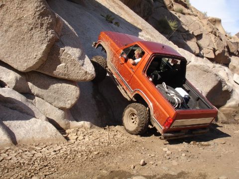 1984 Ford Bronco - An orange off-road vehicle navigates a rocky incline, with visible text from Supermotors.net on the lower right.