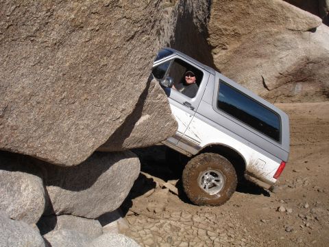 1984 Ford Bronco - A driver maneuvers a Ford Bronco through a challenging rocky terrain, with large boulders on either side.