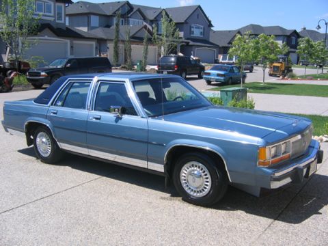 1988 Ford Crown Victoria LTD - A blue sedan with a vinyl roof is parked in a driveway with houses in the background.