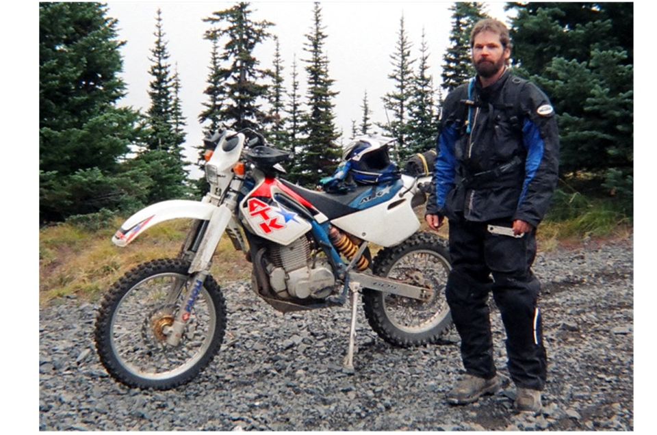 Man in riding gear stands next to a white dirt bike with red and blue graphics. Trees and gravel are in the background