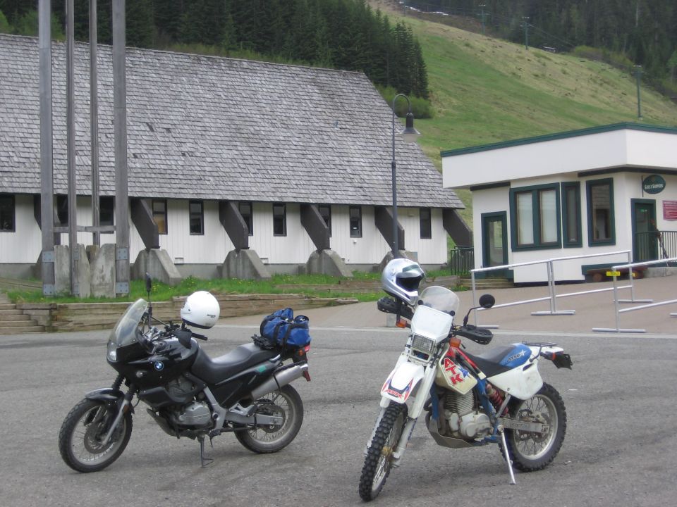 Black BMW F650 motorcycle parked next to a white dirt bike, with a building and grassy hill in the background