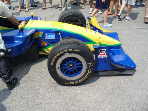 1995 Ford Taurus - A race car with blue and yellow livery is parked on asphalt, with spectators in the background.
