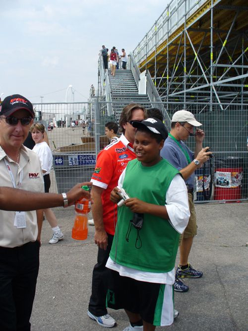 Man in PKV shirt hands drink to kid in green vest, race track stands in background