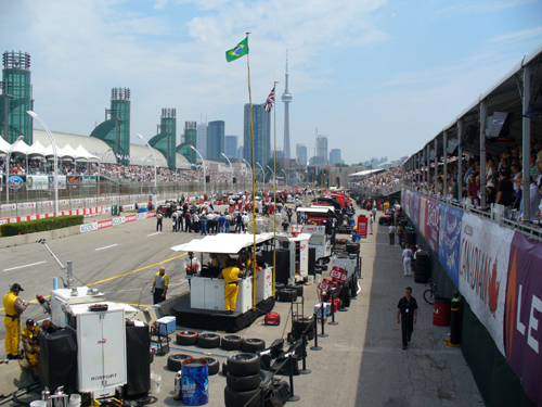 Race track pit lane with tents, tires, and city skyline in background. Brazilian and Canadian flags visible