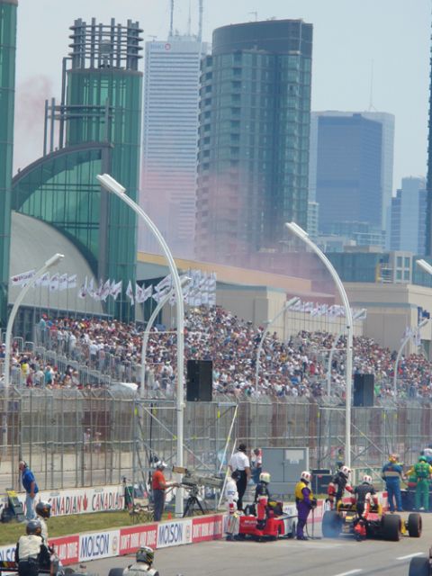 1995 Ford Taurus - Race cars are lined up on a track with a large crowd watching in the stands, set against a city skyline.