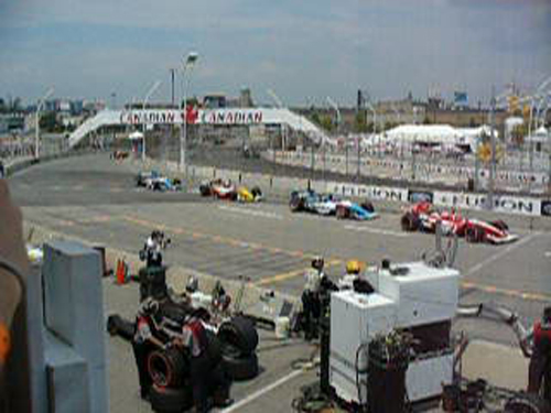 Race cars on a track with pit crew and Canadian banners overhead