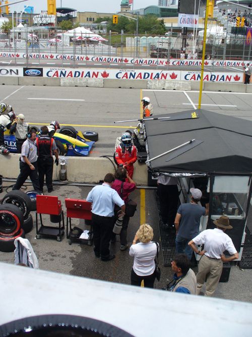 Race car pit crew and media gathered around a car in a pit stop area. A driver in a red suit stands near a tent