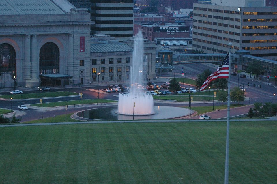 Cityscape with a large fountain, buildings, American flag, and several cars parked and driving
