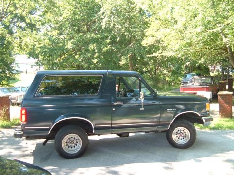 1988 Ford Bronco - A dark green SUV is parked on a paved surface with trees and other vehicles in the background.