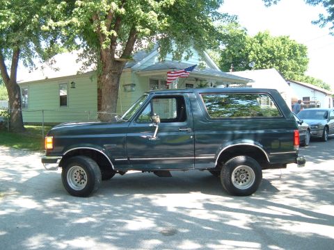 1988 Ford Bronco - A dark green 1988 Ford Bronco is parked on a residential street, with another vehicle visible in the background.