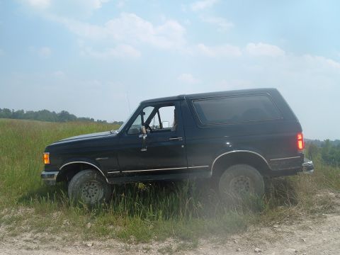 1988 Ford Bronco - A black SUV navigates a grassy, off-road trail under a partly cloudy sky.
