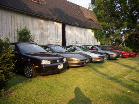 1996 Ford Probe - A collection of coupes and a hatchback are lined up on a grassy lawn next to a weathered barn.