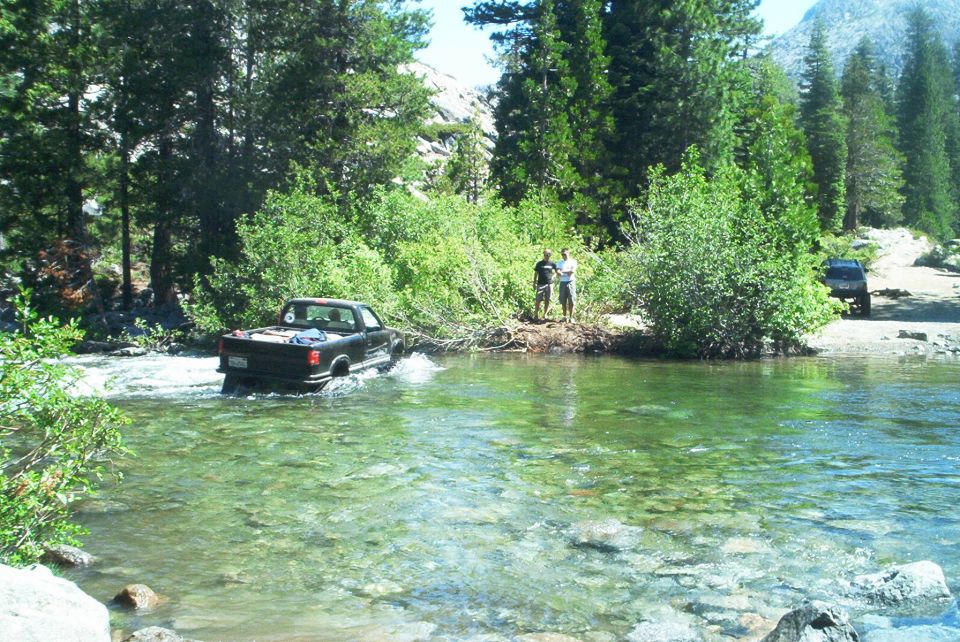 Black pickup fording a clear rocky mountain river with onlookers on the far bank