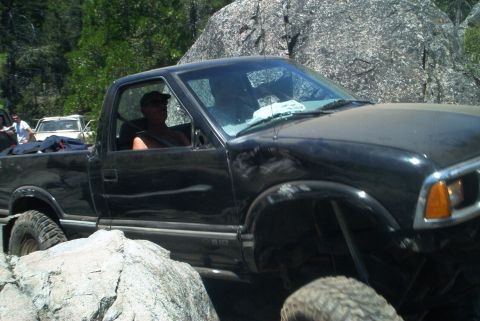 1966 Jeep CJ5 - A black pickup truck is shown off-roading over large rocks, with another vehicle visible behind it.