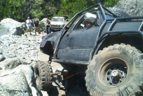 1966 Jeep CJ5 - This heavily modified off-road vehicle navigates a challenging rocky trail, with other vehicles and people visible in the background.