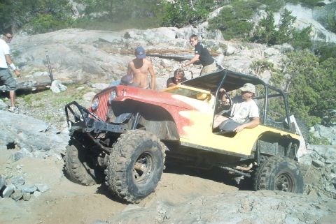 1966 Jeep CJ5 - A heavily modified Jeep CJ-5 is shown off-roading on a steep, rocky incline with several people present.