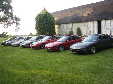 1996 Ford Probe - Seven Ford Probe coupes in black, silver, red and dark green lined up nose-out on mown grass beside a weathered barn with peeling white sid…