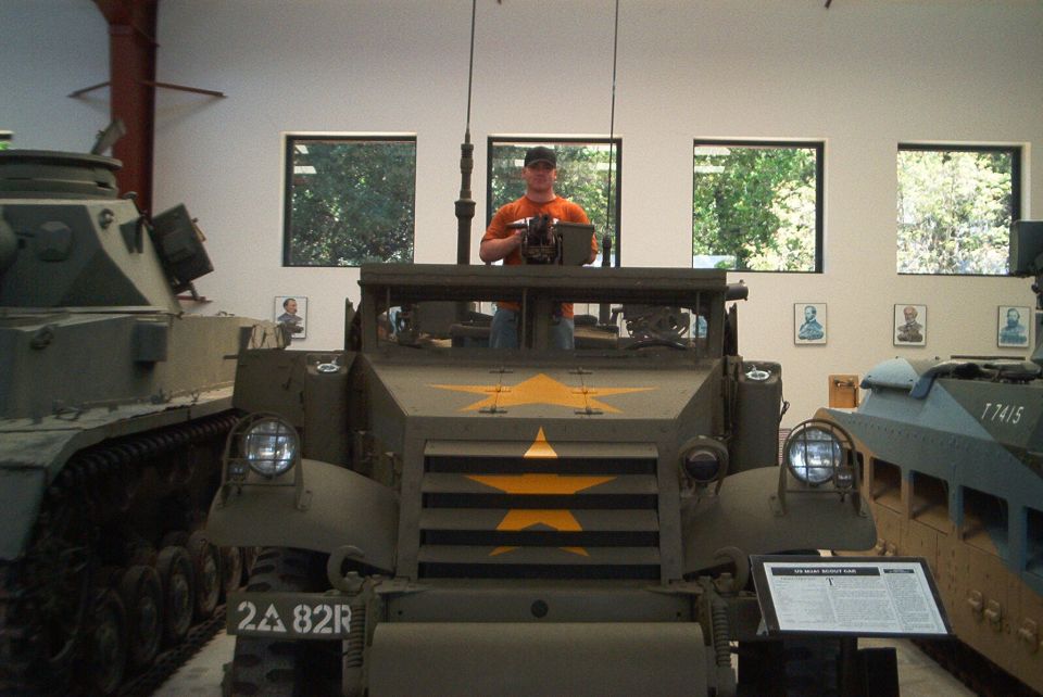 Front view of olive drab armored vehicle with yellow stars, man behind machine gun, other military vehicles nearby