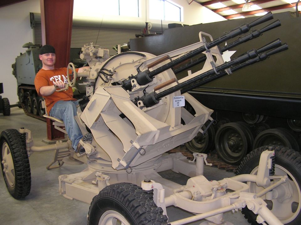 Person sits in the operator's seat of a quad-barreled anti-aircraft gun mount. Another tracked military vehicle is visible behind it