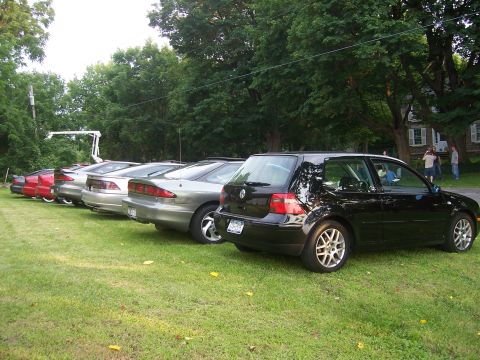 1996 Ford Probe - A line of cars, including a black Volkswagen Golf and several silver Ford Probes, are parked on a grassy area near trees and under trees.